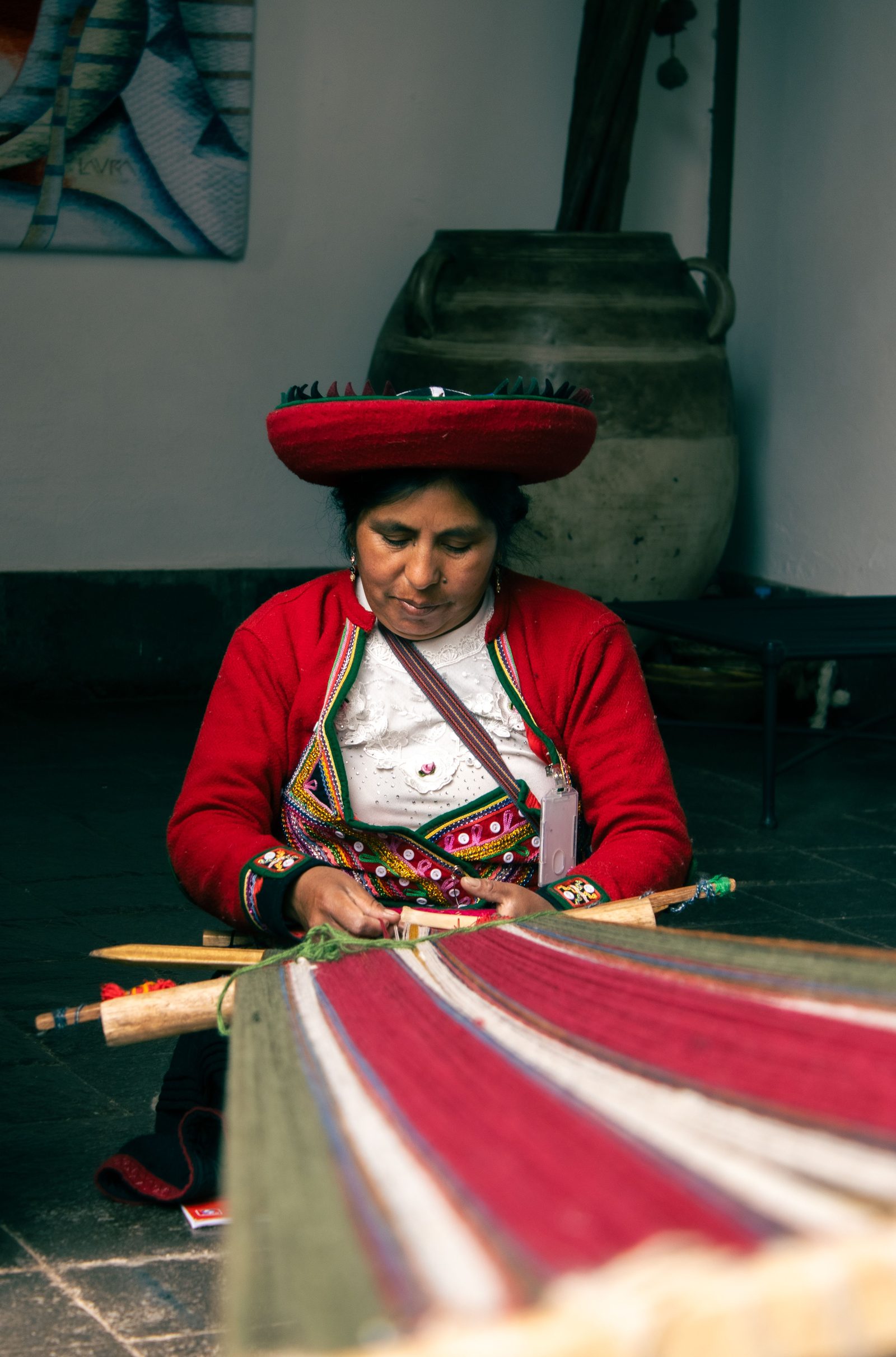 A weaver works an indoor backstrap loom, red hat and vest