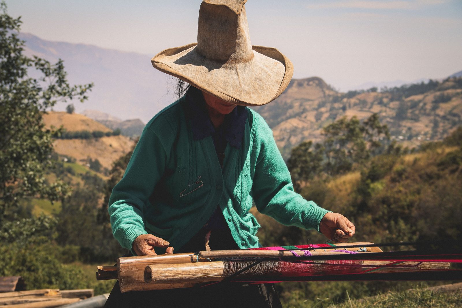 A weaver works a backstrap loom outdoors, Andean foothills in the background